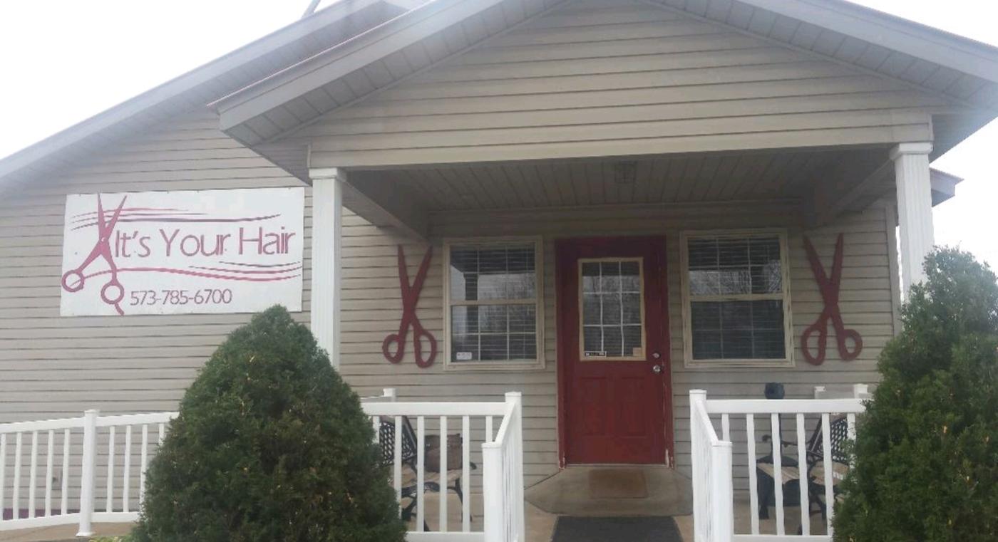 The salon: a cottage with cream siding, a covered porch, white railings and a red front door, with a painted sign and two oversized scissors mounted on the wall either side of the door.